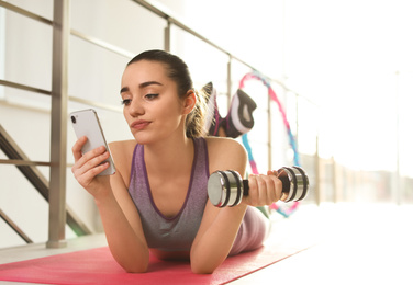 Photo of Lazy young woman with dumbbell and smartphone on yoga mat indoors