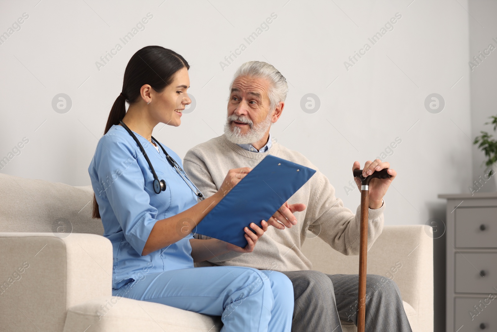 Photo of Smiling nurse with clipboard assisting elderly patient on sofa in hospital