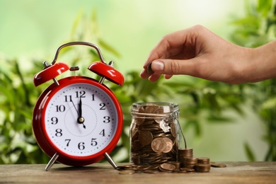Woman putting coin into glass jar and red alarm clock on wooden table, closeup. Money savings