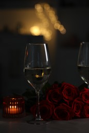 Glasses of white wine, burning candle and rose flowers on grey table against blurred lights. Romantic atmosphere