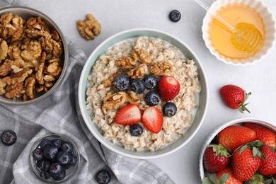 Photo of Tasty oatmeal with strawberries, blueberries and walnuts surrounded by ingredients on grey table, flat lay