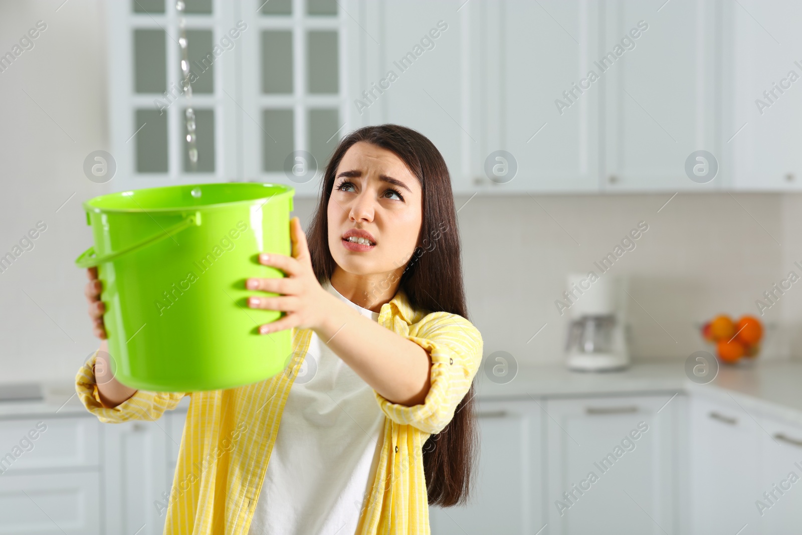 Photo of Young woman collecting leaking water from ceiling in kitchen, space for text. Time to call roof repair service