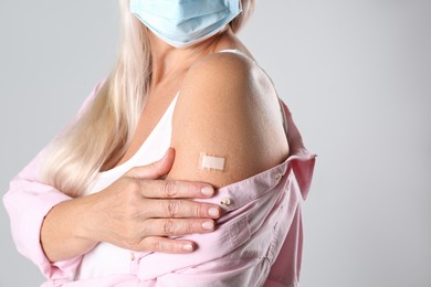 Photo of Mature woman in protective mask showing arm with bandage after vaccination on light background, closeup