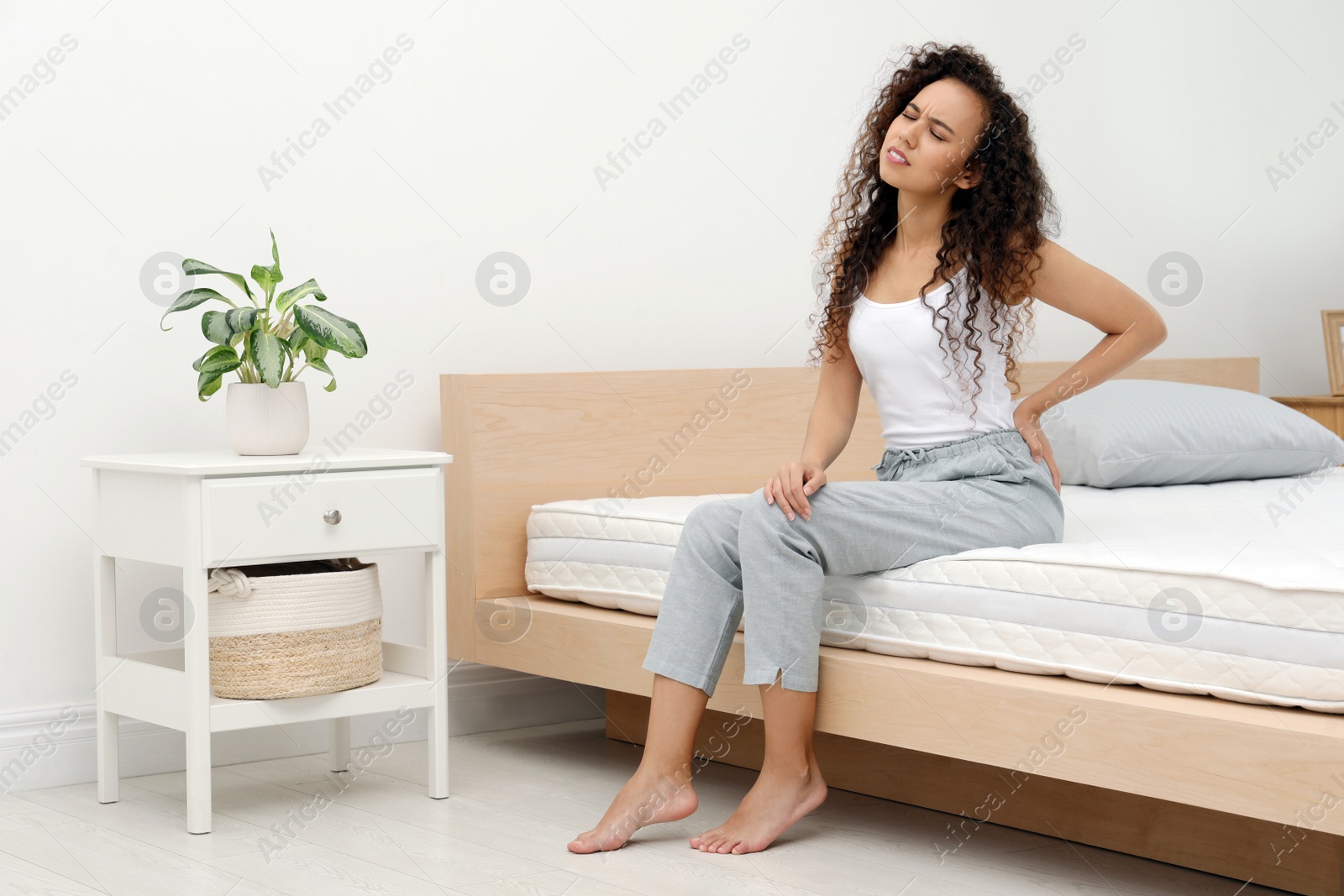 Photo of Young African American woman suffering from back pain after sleeping on uncomfortable mattress in bedroom