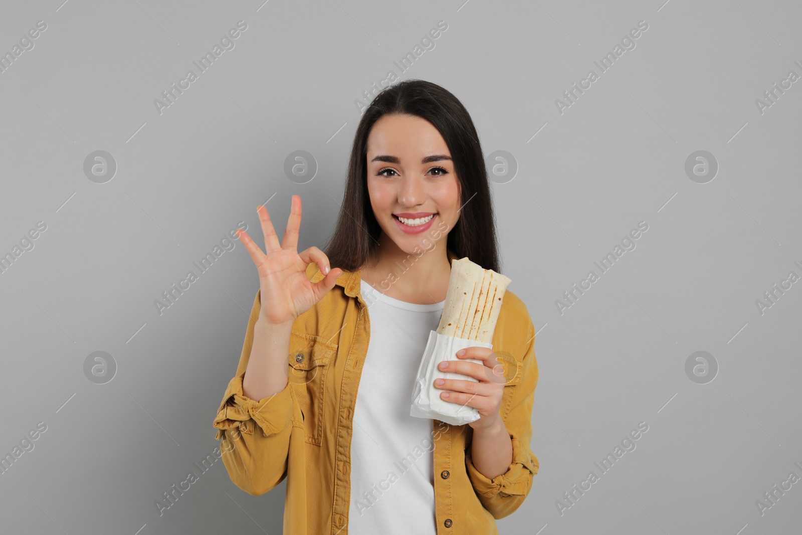 Photo of Happy young woman with tasty shawarma showing okay gesture on grey background