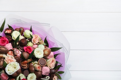 Beautiful bouquet of flowers and chocolate candies on white wooden background, top view. Space for text