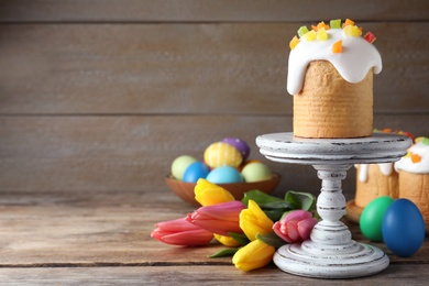 Photo of Stand with traditional Easter cake, tulips and colorful eggs on wooden table. Space for text