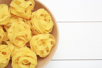 Photo of Raw tagliatelle pasta in bowl on white wooden table, top view. Space for text