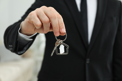 Photo of Real estate agent holding house key with trinket indoors, closeup