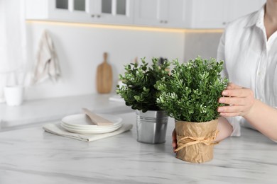 Woman near white marble table with different artificial potted herbs in kitchen, closeup. Space for text