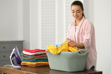 Photo of Young woman with basket full of clean laundry at wooden table indoors