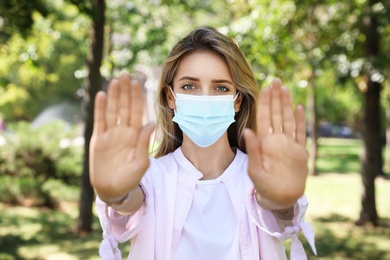 Photo of Woman in protective face mask showing stop gesture in park. Prevent spreading of coronavirus