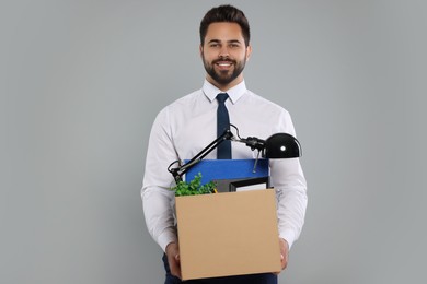 Happy unemployed man with box of personal office belongings on light grey background