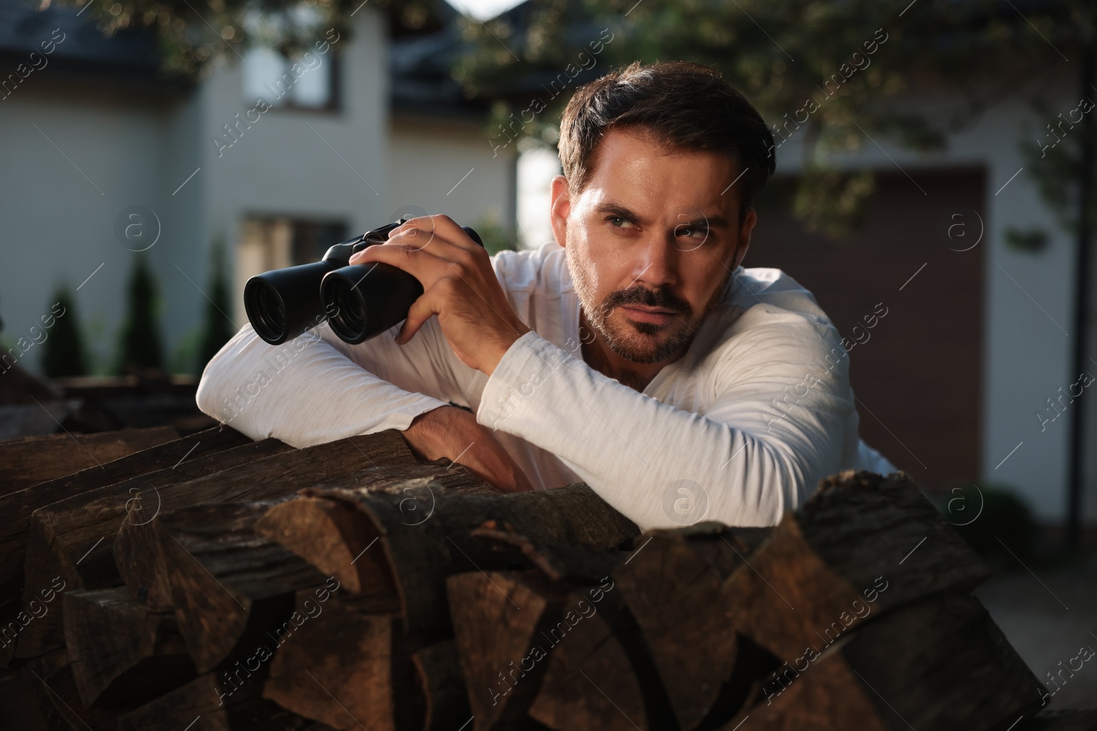 Photo of Concept of private life. Curious man with binoculars spying on neighbours over firewood outdoors