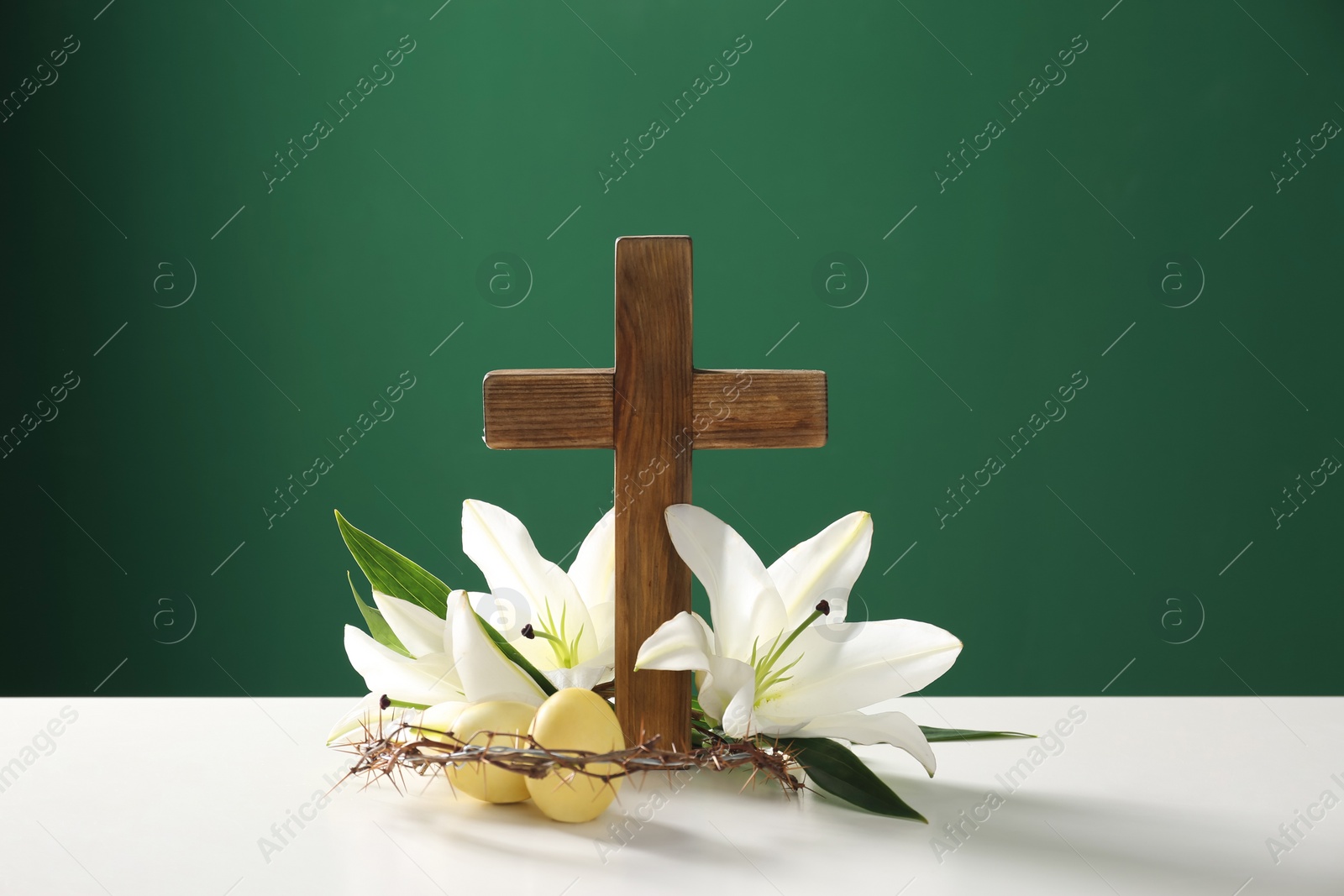 Photo of Wooden cross, crown of thorns, Easter eggs and blossom lilies on table against color background