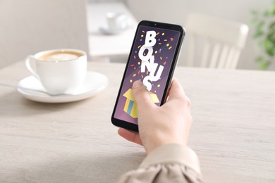 Image of Bonus gaining. Woman holding smartphone at white wooden table, closeup. Illustration of open gift box, word and confetti on device screen