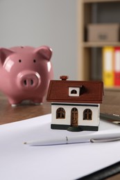 Photo of House model, piggy bank, clipboard and pen on wooden table indoors, selective focus