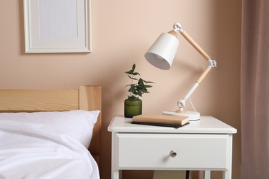 Photo of Stylish modern desk lamp, books and plant on white nightstand in bedroom