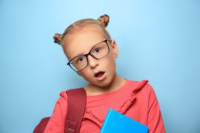 Photo of Cute little girl with glasses, backpack and textbook on light blue background