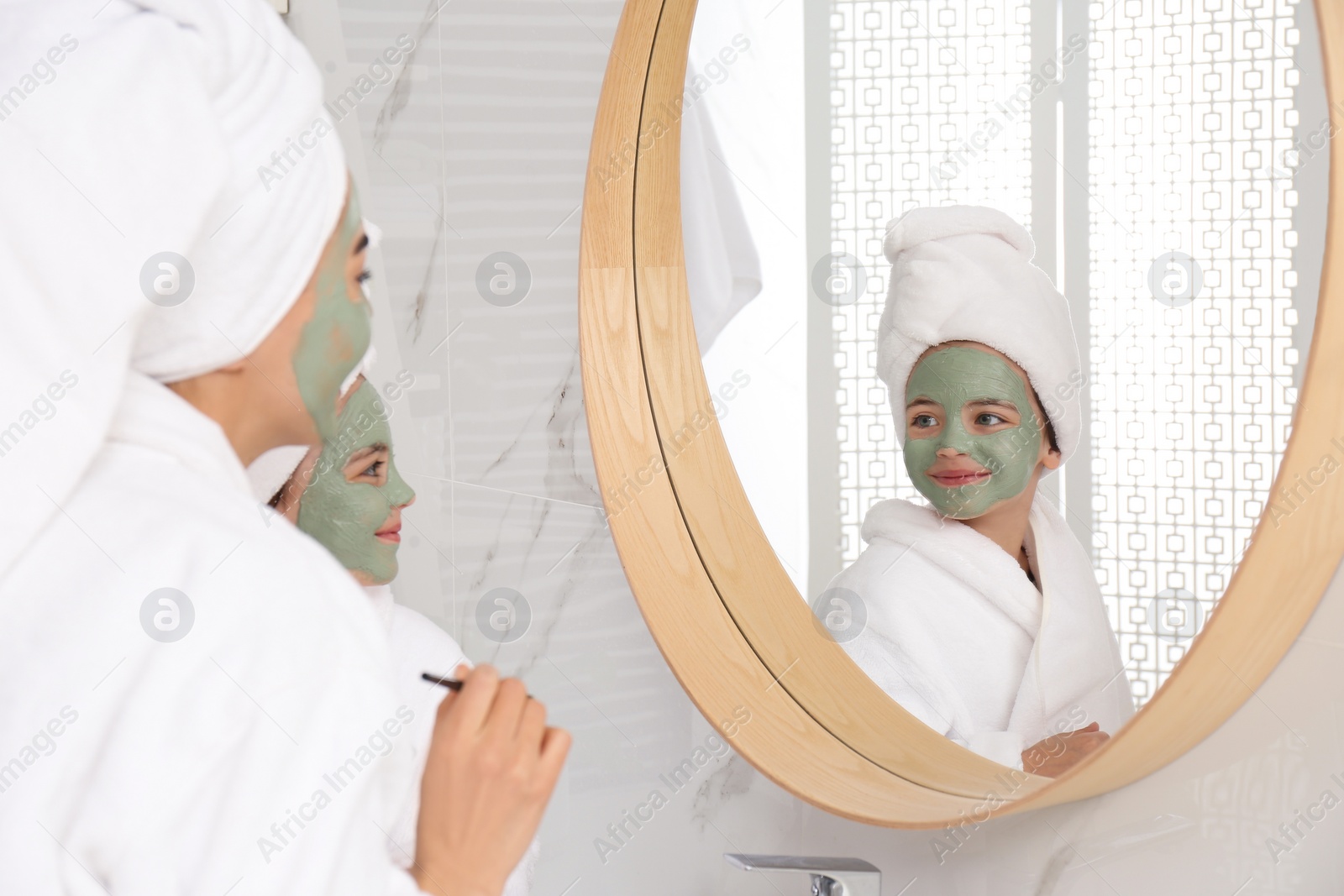 Photo of Young mother and her daughter with facial masks near mirror in bathroom