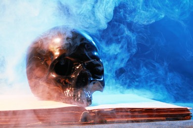 Black human skull, old book and smoke on stone surface against color background