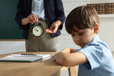 Photo of Teacher scolding pupil for being late in classroom, focus on alarm clock