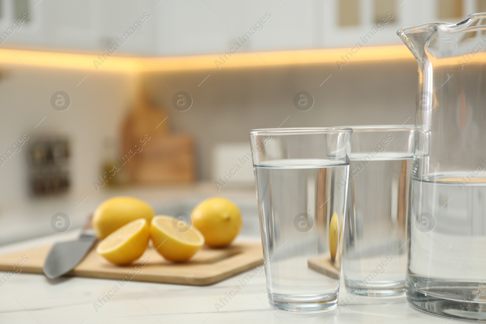 Photo of Jug, glasses with clear water and lemons on white table in kitchen, closeup