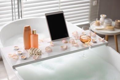 Photo of Wooden tray with tablet, wine, toiletries and flower petals on bathtub in bathroom