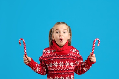 Surprised little girl in knitted Christmas sweater holding candy canes on blue background