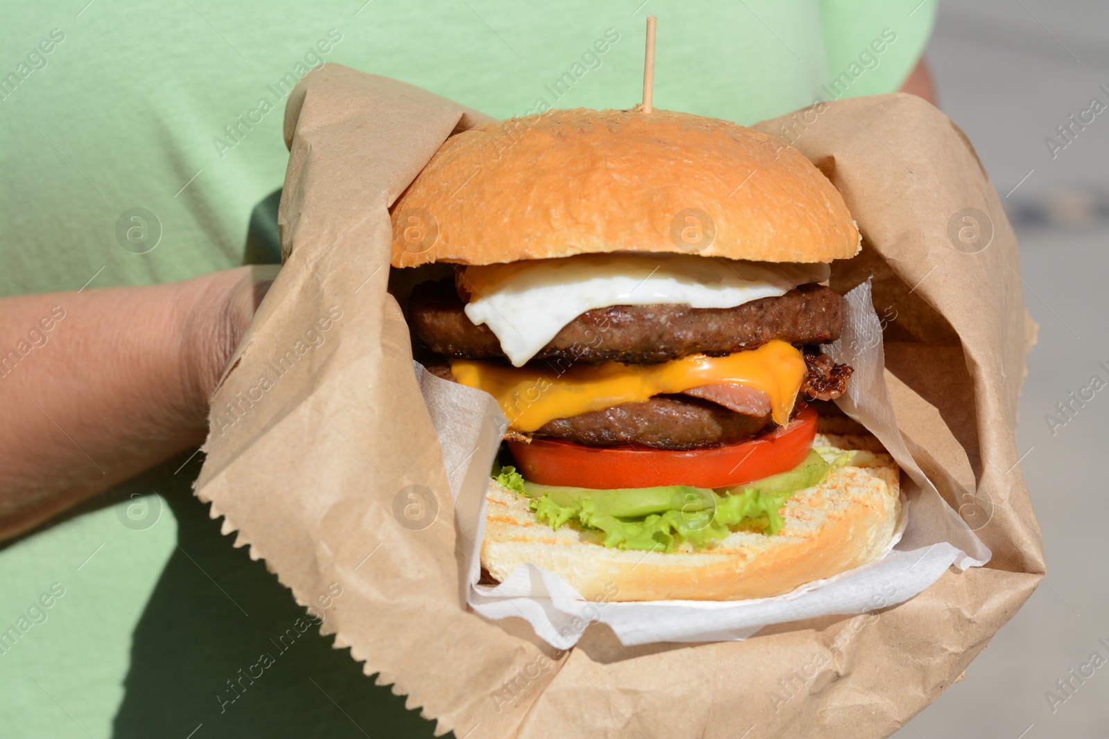 Photo of Woman holding delicious burger in paper wrap on blurred background, closeup