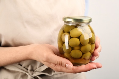 Photo of Woman holding glass jar of pickled olives on light background, closeup
