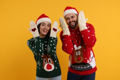 Photo of Young couple in Christmas sweaters, Santa hats and knitted mittens on orange background