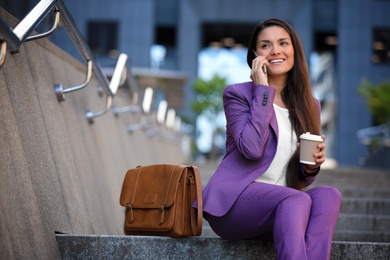 Photo of Beautiful businesswoman with cup of coffee talking on phone outdoors