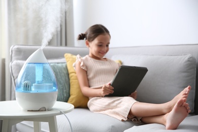 Photo of Little girl using tablet in room with modern air humidifier
