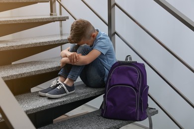 Upset little boy with backpack sitting on stairs indoors