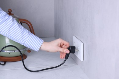 Photo of Woman plugging electric kettle into power socket at white table indoors, closeup