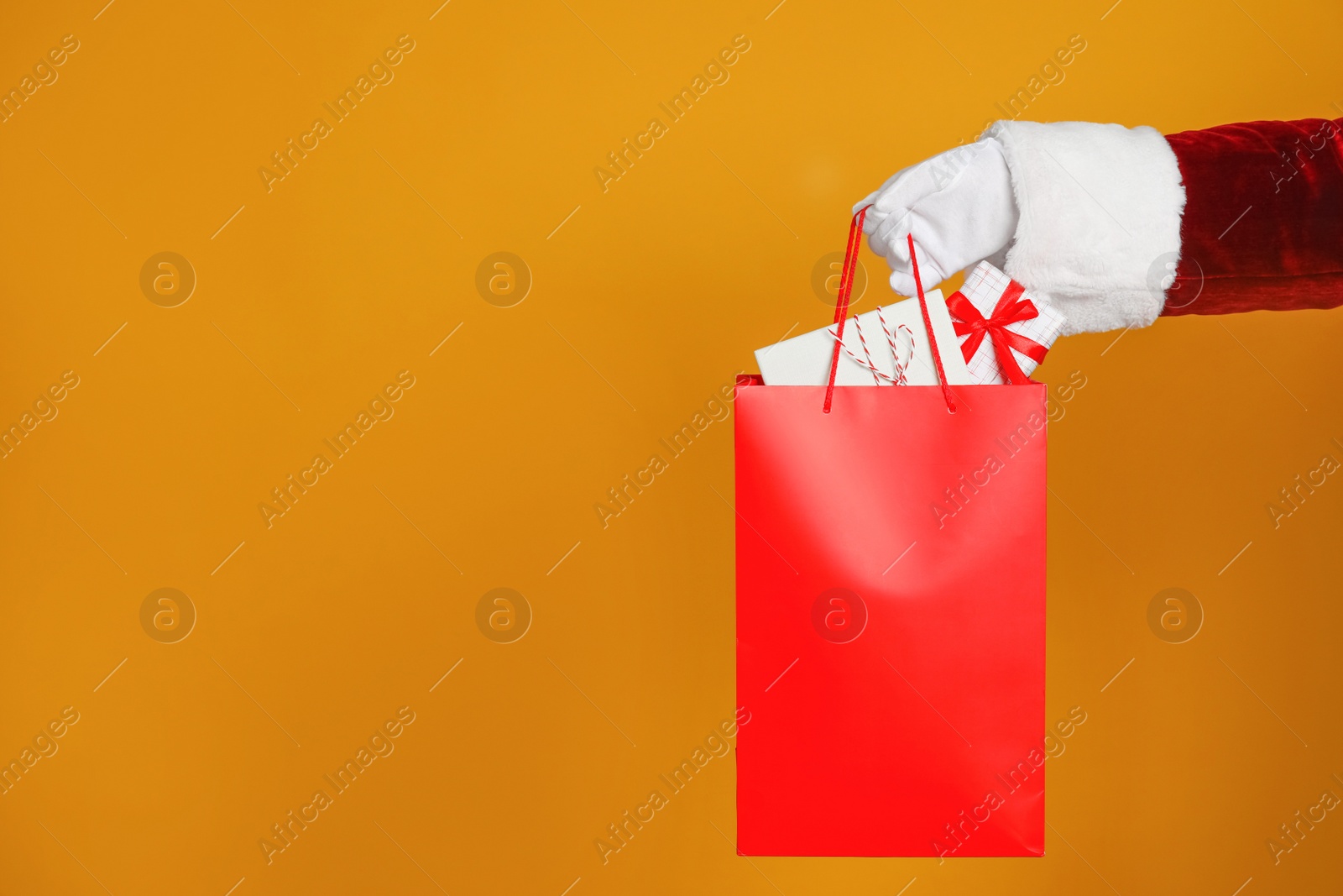 Photo of Santa holding paper bag with gift boxes on orange background, closeup. Space for text