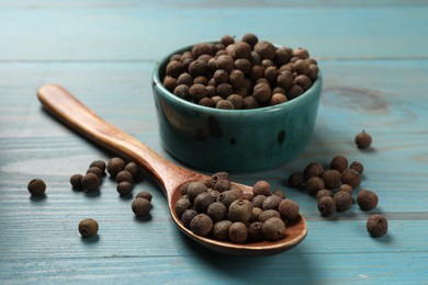 Photo of Aromatic allspice pepper grains in bowl and spoon on light blue wooden table, closeup