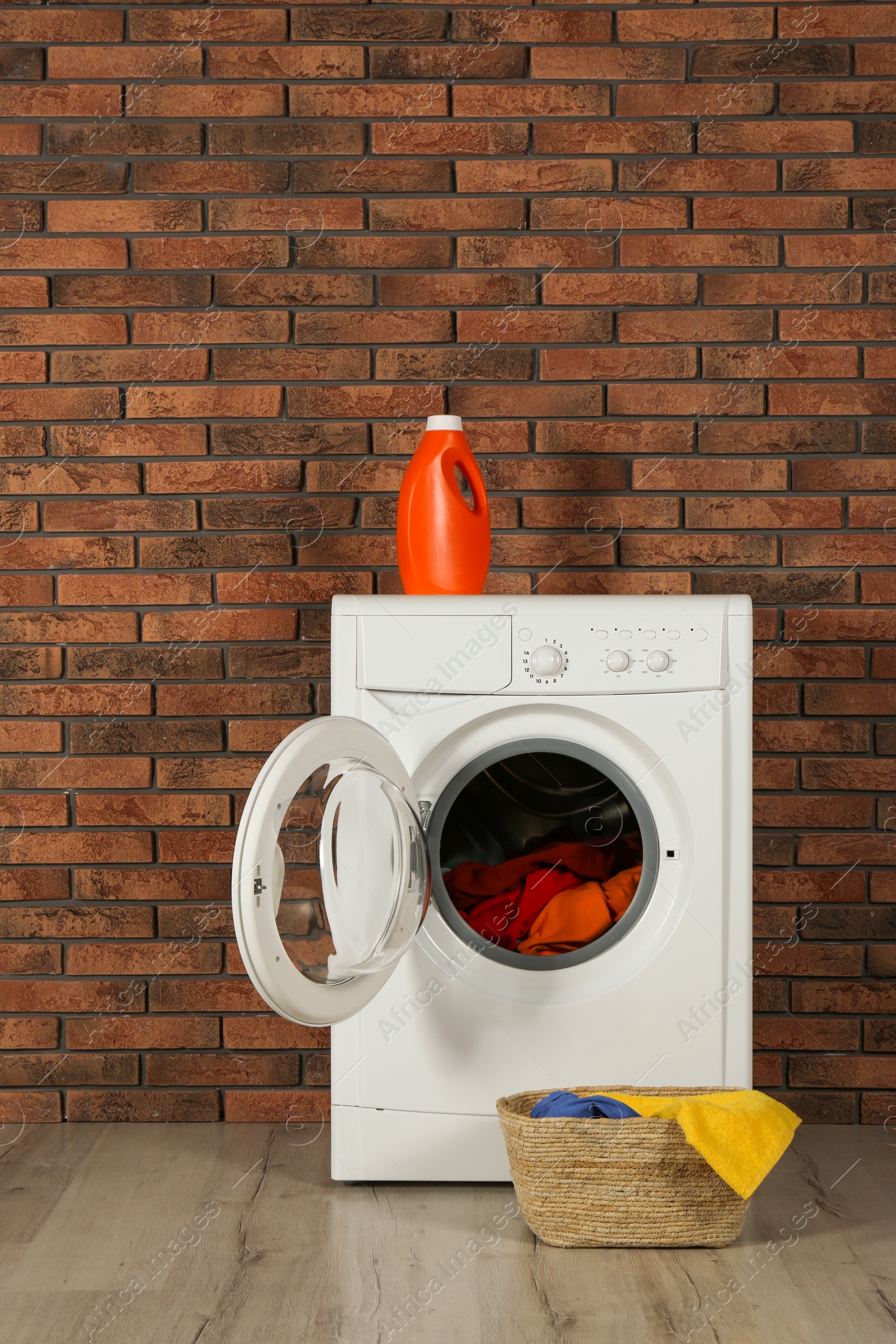 Photo of Modern washing machine with laundry, detergent and wicker basket near brick wall