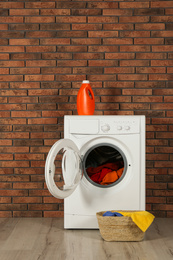 Modern washing machine with laundry, detergent and wicker basket near brick wall