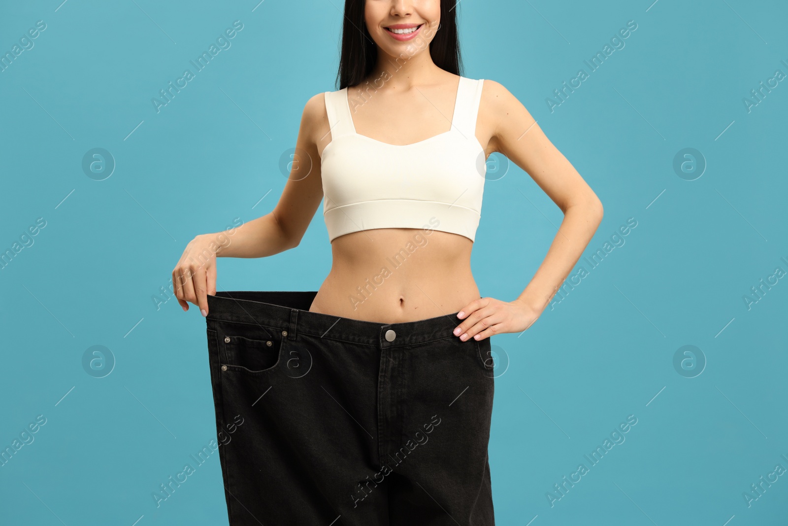 Photo of Happy young woman wearing big jeans after weight loss on turquoise background, closeup