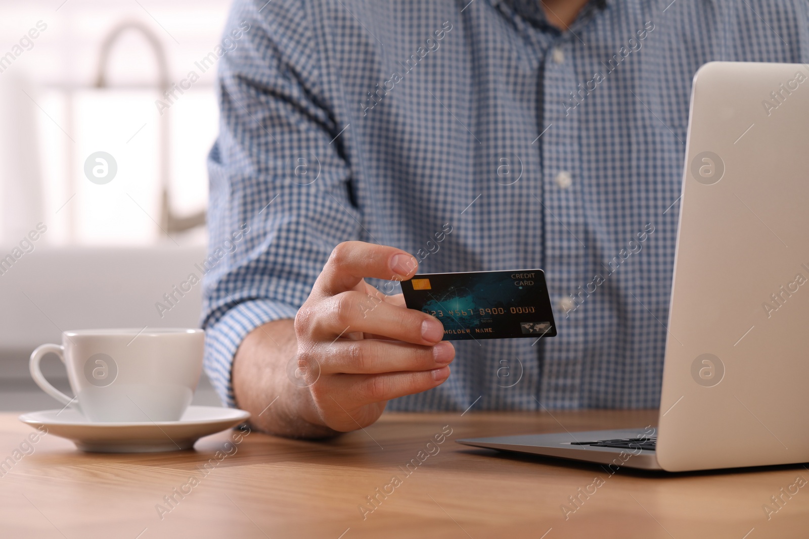 Photo of Man using laptop and credit card for online payment at table in kitchen, closeup