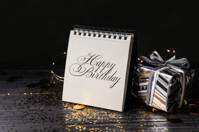 Photo of Notepad with greeting HAPPY BIRTHDAY and gift on wooden table