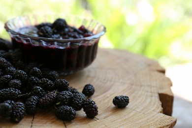 Delicious ripe black mulberries and bowl of sweet jam on wood stump outdoors, closeup. Space for text