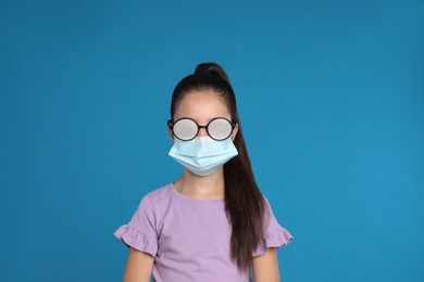 Photo of Little girl with foggy glasses caused by wearing medical face mask on blue background. Protective measure during coronavirus pandemic
