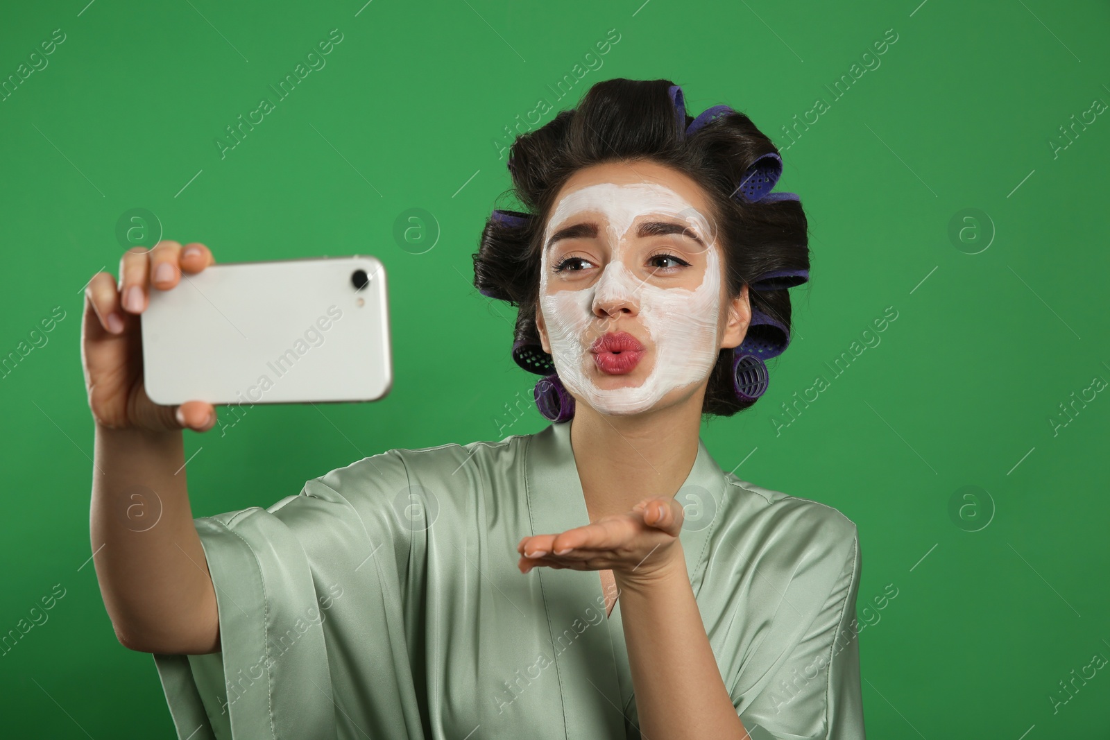 Photo of Young woman in silk bathrobe with hair curlers and facial mask taking selfie on green background