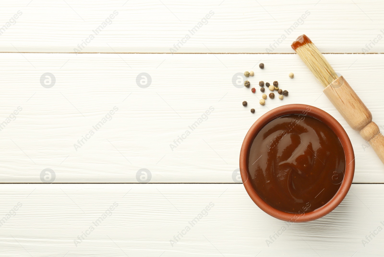Photo of Tasty barbeque sauce in bowl, brush and peppercorns on white wooden table, top view. Space for text