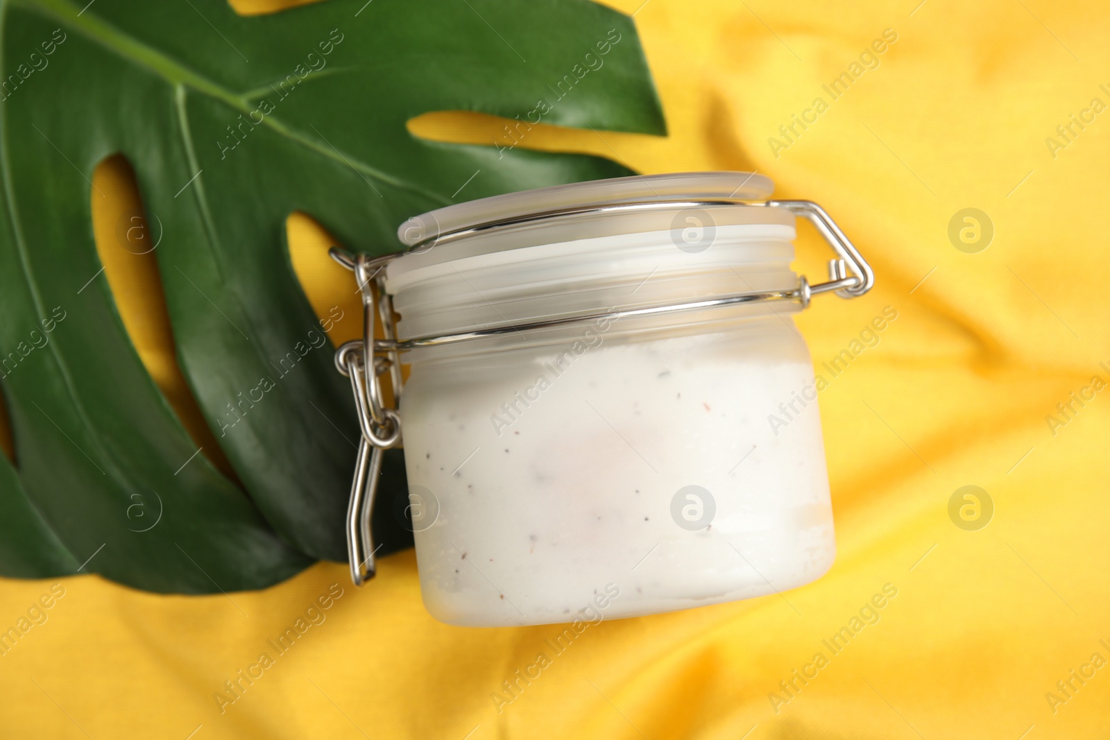 Photo of Jar of salt scrub and palm leaf on yellow fabric, flat lay