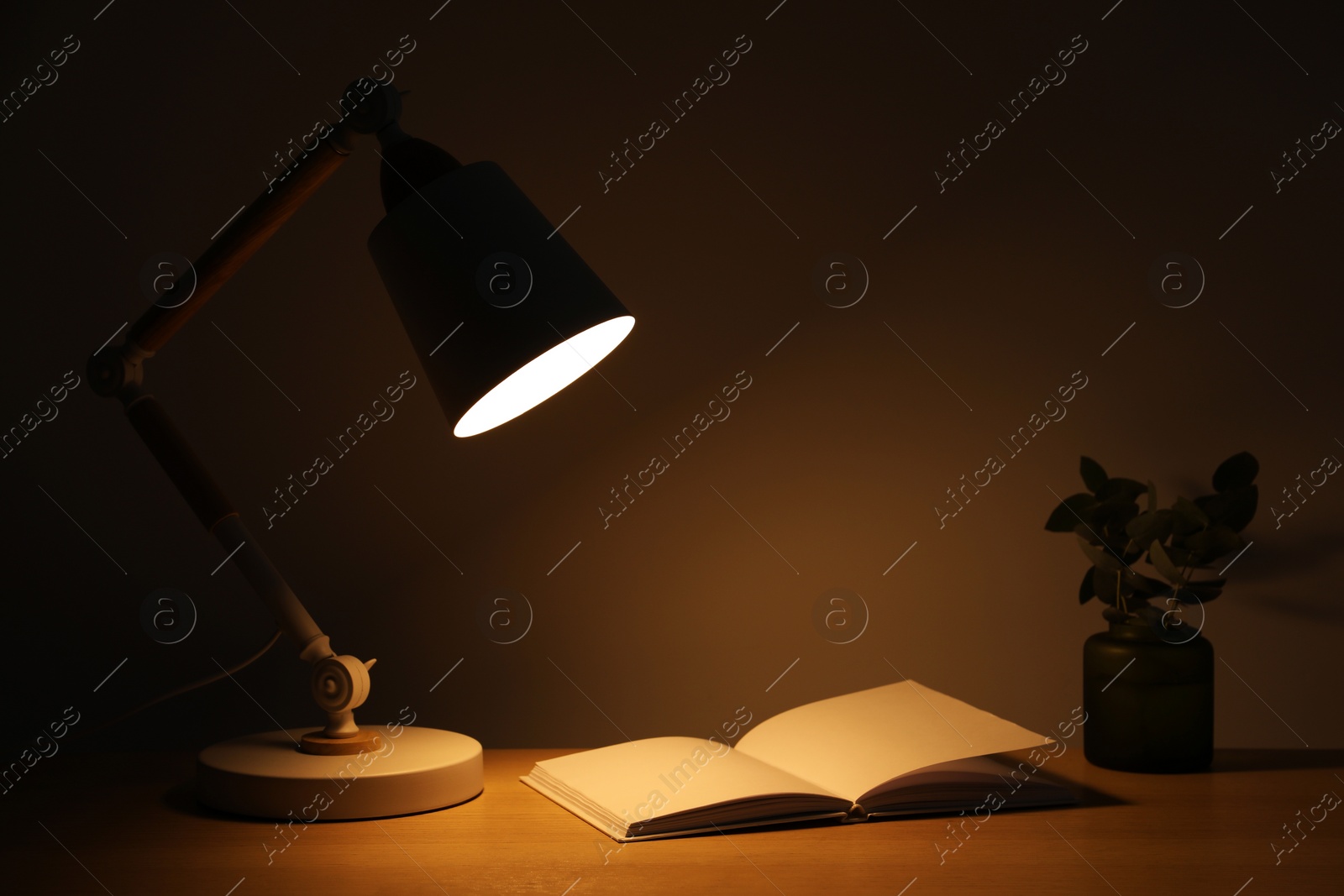 Photo of Stylish modern desk lamp, open book and plant on wooden table near wall in dark room