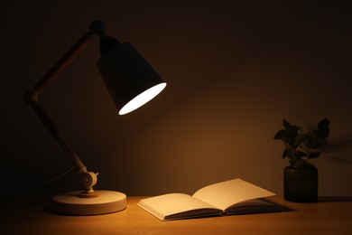 Photo of Stylish modern desk lamp, open book and plant on wooden table near wall in dark room